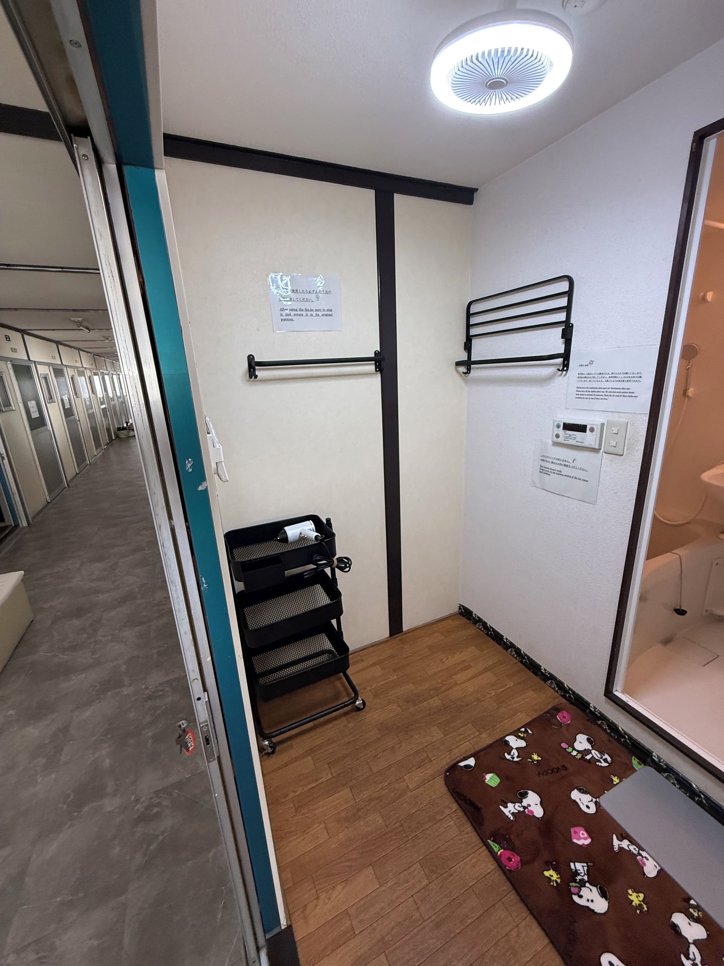 A hotel room with a wooden floor, a black metal rack, a wall-mounted towel rack, and a door leading to a bathroom.