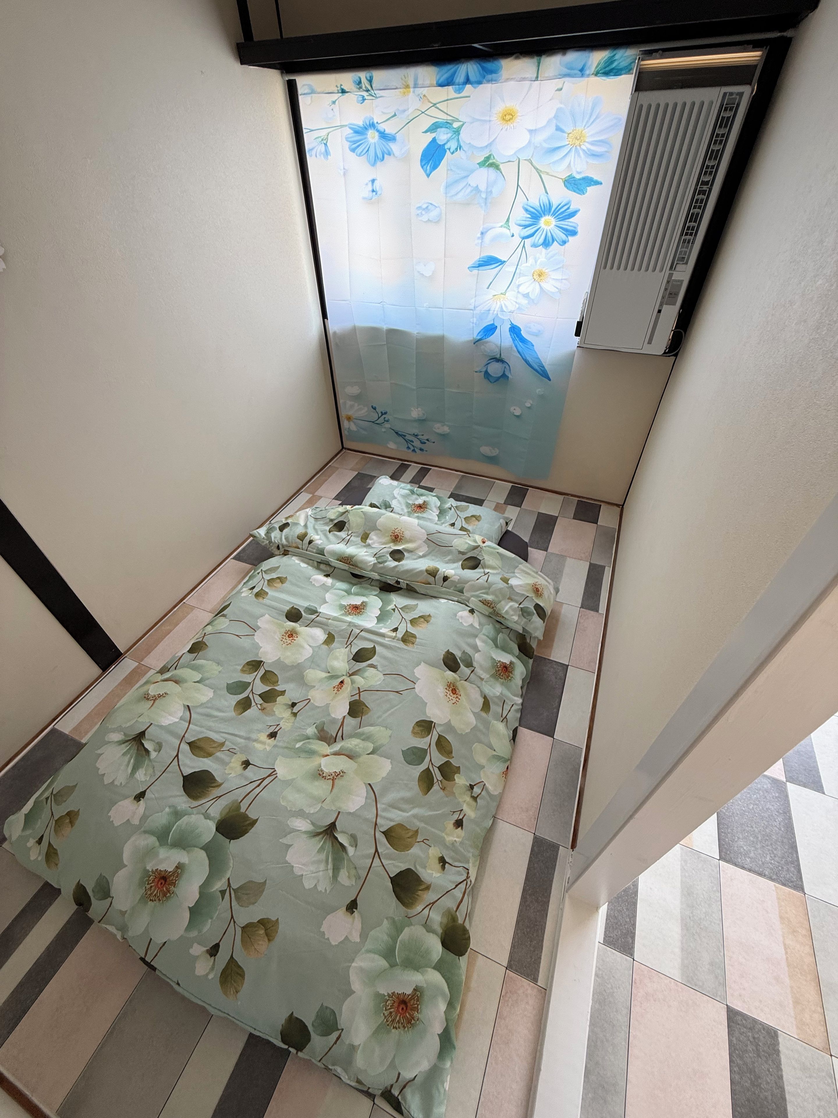 A small room with a bed, a window with a floral curtain, and a tiled floor.