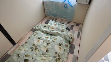 A small room with a bed, a window with a floral curtain, and a tiled floor.
