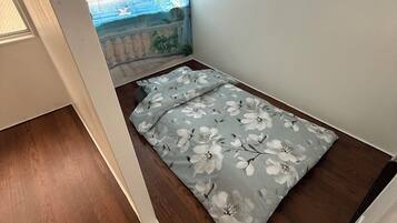 A small room with a bed, a window with a view of palm trees and the sea, and a wooden floor.