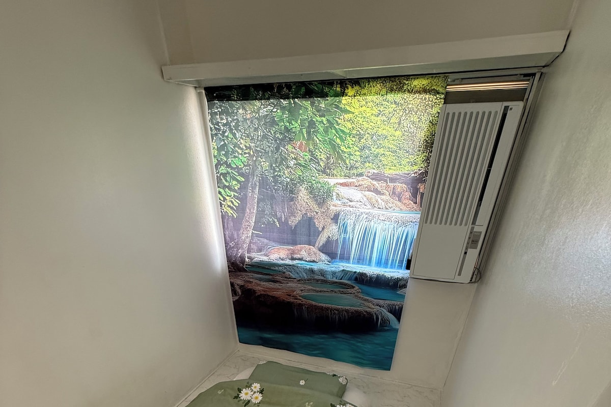 A small, narrow room with a bed featuring a floral bedspread and pillows, a window with a view of a waterfall, and a wall-mounted air conditioning unit.