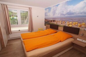 Ein Schlafzimmer mit einem Bett, einem Fenster mit Vorhängen, einem Wandbild mit Strandmotiv und einem kleinen Tisch.