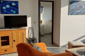 A living room with a television, wooden cabinet, orange sofa, and a view into the kitchen. - Holiday apartment in Damp with swimming pool (Damp)