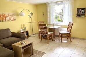 Ein Wohnzimmer mit einer Couch, einem Couchtisch und zwei Holzstühlen. Ein Fenster mit Vorhängen, eine Lampe und ein Gemälde sind an der Wand zu sehen.