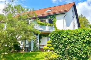 Een twee verdiepingen tellend huis met een dak van rode tegels, een balkon met klimplanten en een verzorgde tuin.