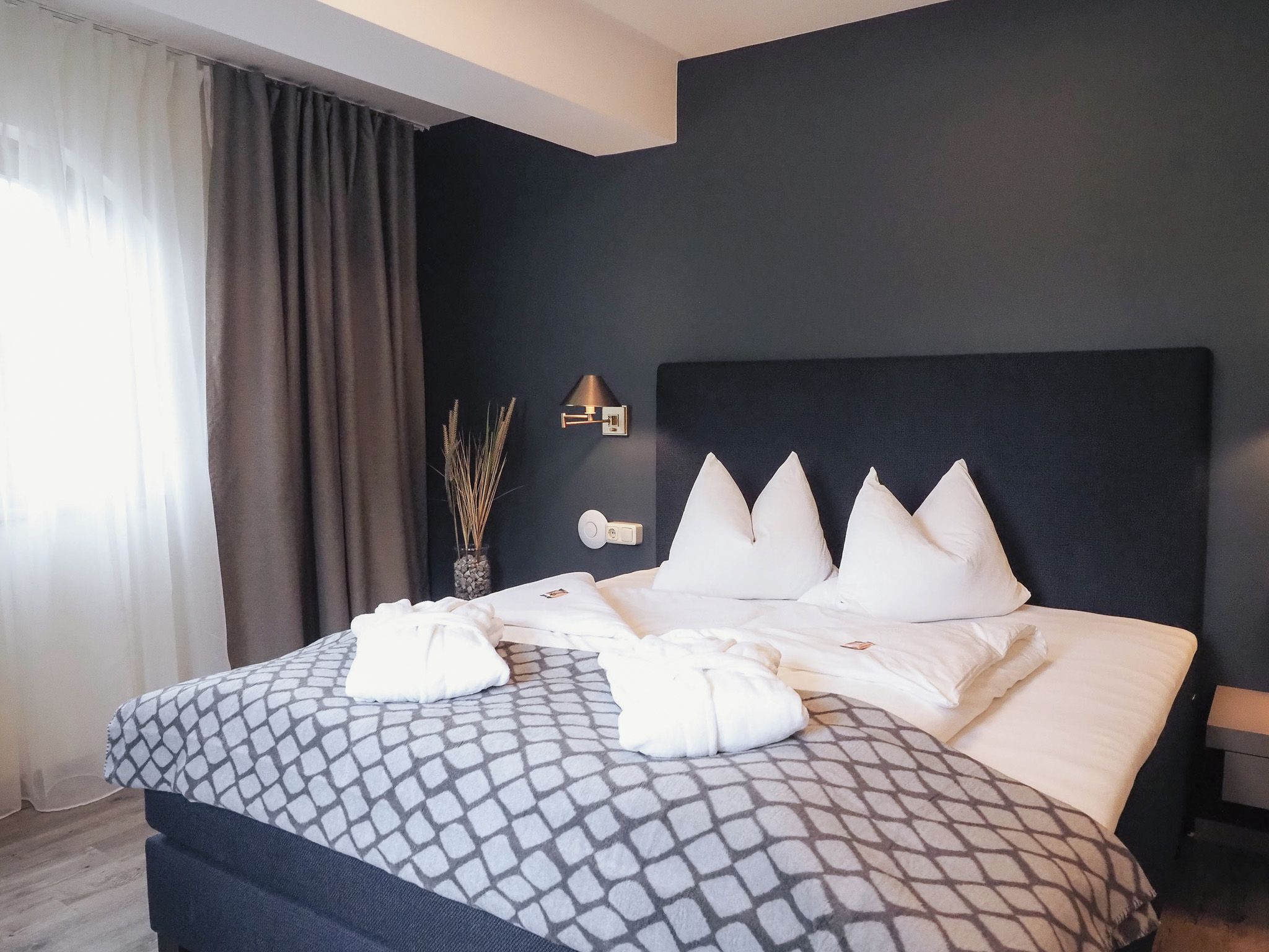 Ein modernes Hotelzimmer mit einem großen Bett, einem dunklen Kopfteil und einem Nachttisch mit einer Lampe.