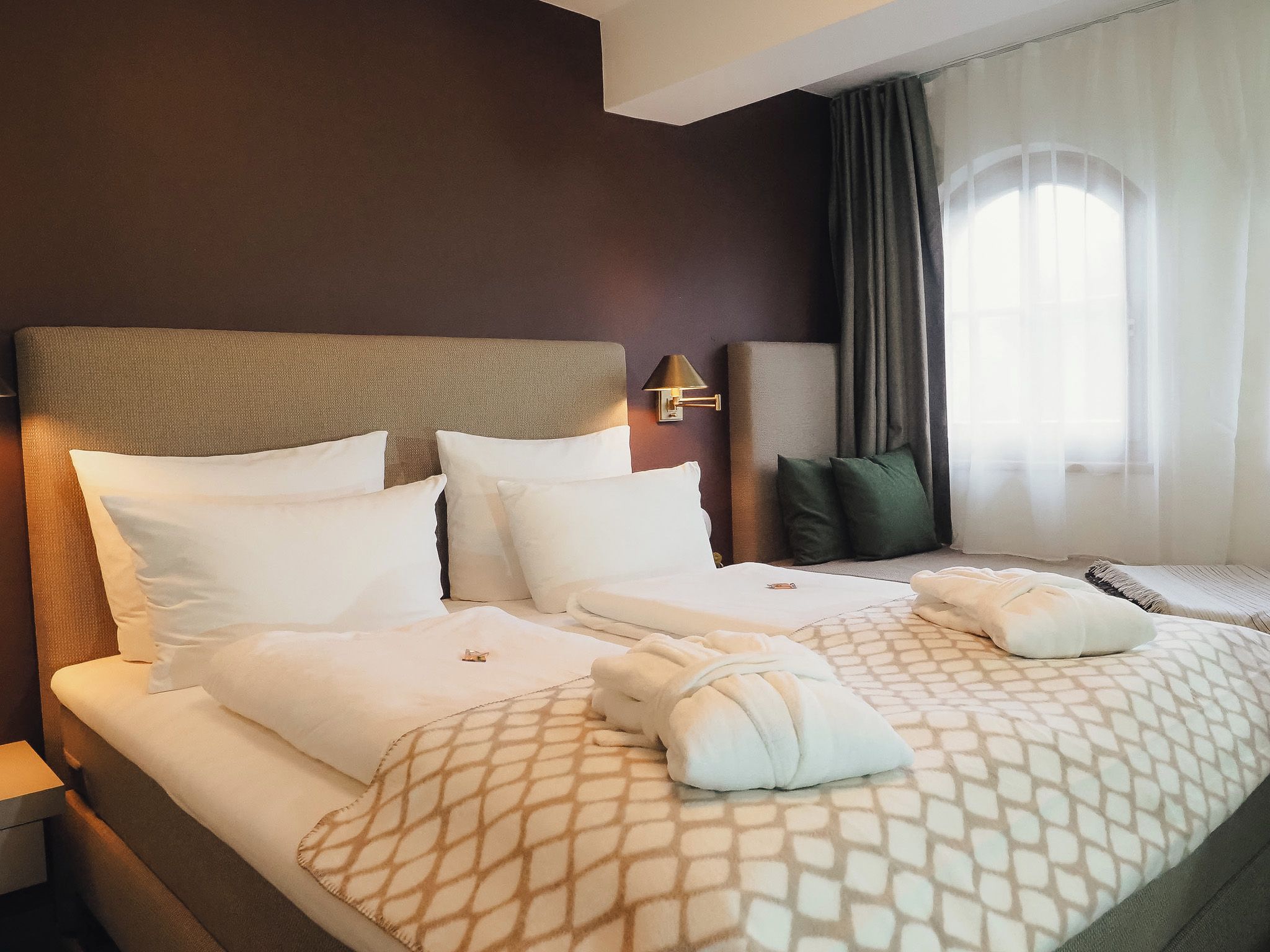 Una camera d'albergo con un letto grande, due cuscini, una testiera, una lampada da comodino, una finestra con tende e un comodino.