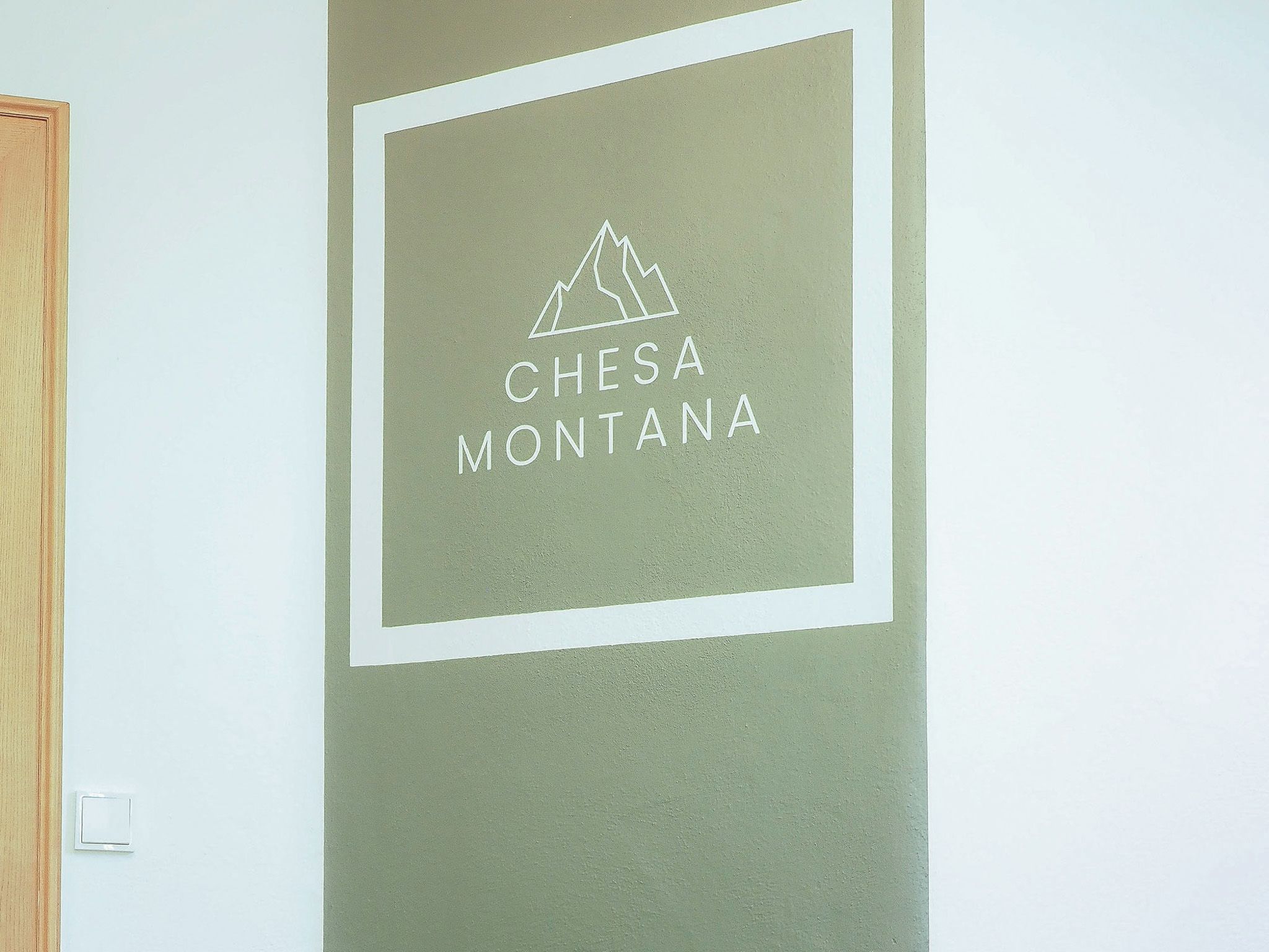 Eine Wand mit einem gerahmten Schild, auf dem 'CHESA MONTANA' steht, und einer Bergsilhouette.