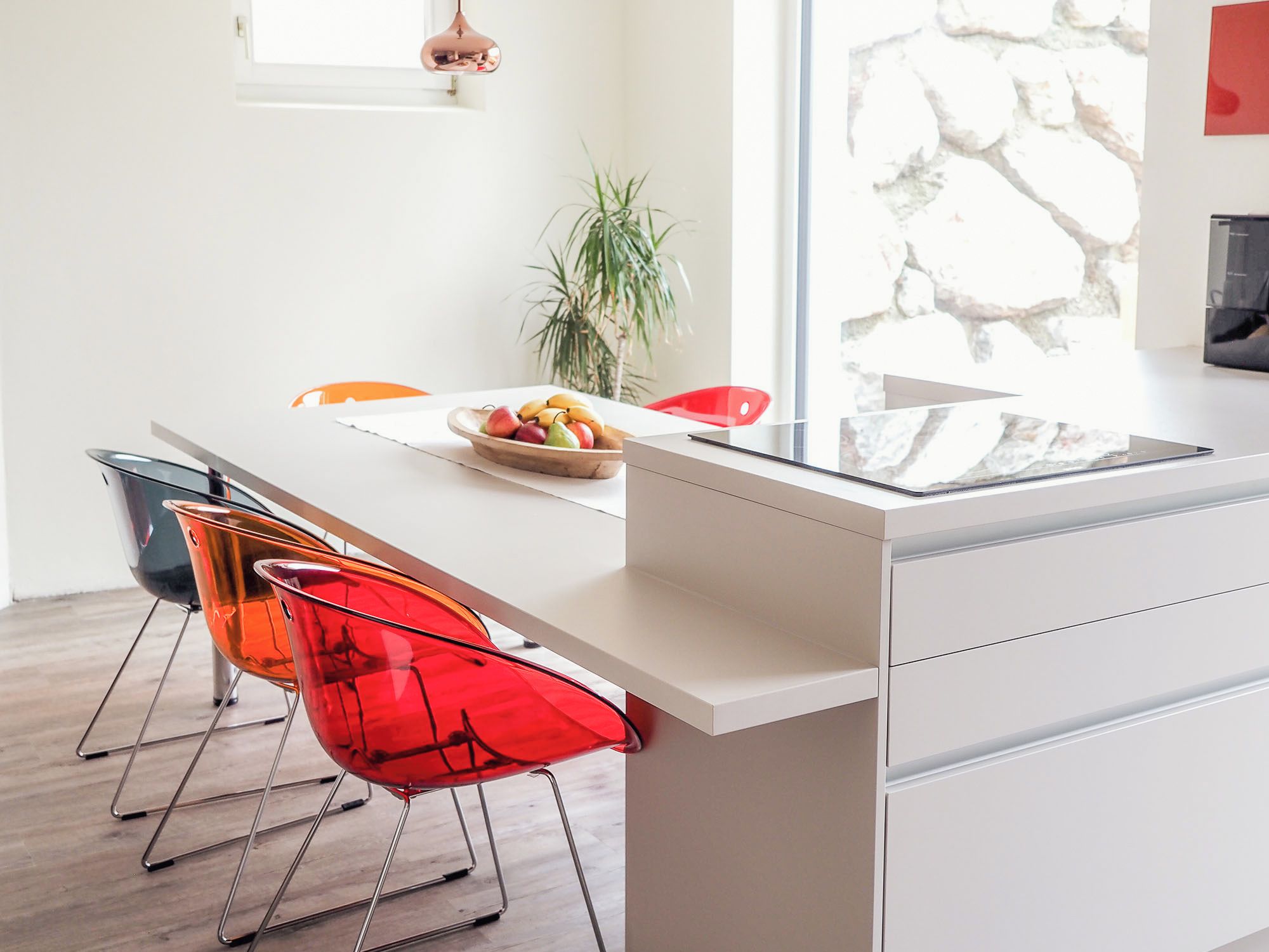 A modern kitchen with a white countertop, a dining area with a red chair, and a potted plant.
