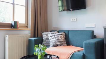 Una sala de estar moderna con un sofá azul, cojines estampados, una mesita redonda con una planta, un televisor montado en la pared y una ventana con cortinas.