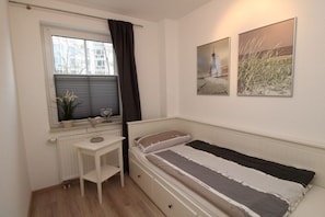 Ein kleines, ordentlich eingerichtetes Schlafzimmer mit einem Einzelbett, einem Nachttisch, einem Heizkörper und einem Fenster mit Vorhang.
