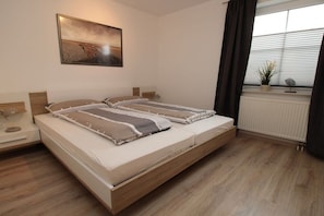 A double bed with a wooden frame, a bedside table with a lamp, a radiator, and a window with curtains. - Holiday apartment with 2 bedrooms (Cuxhaven)