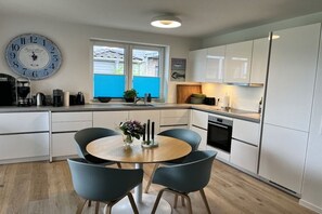 A modern kitchen with white cabinets, a round wooden table with chairs, a large wall clock, and a window with blue shutters. - Holiday home in Gelting with swimming pool (Gelting)