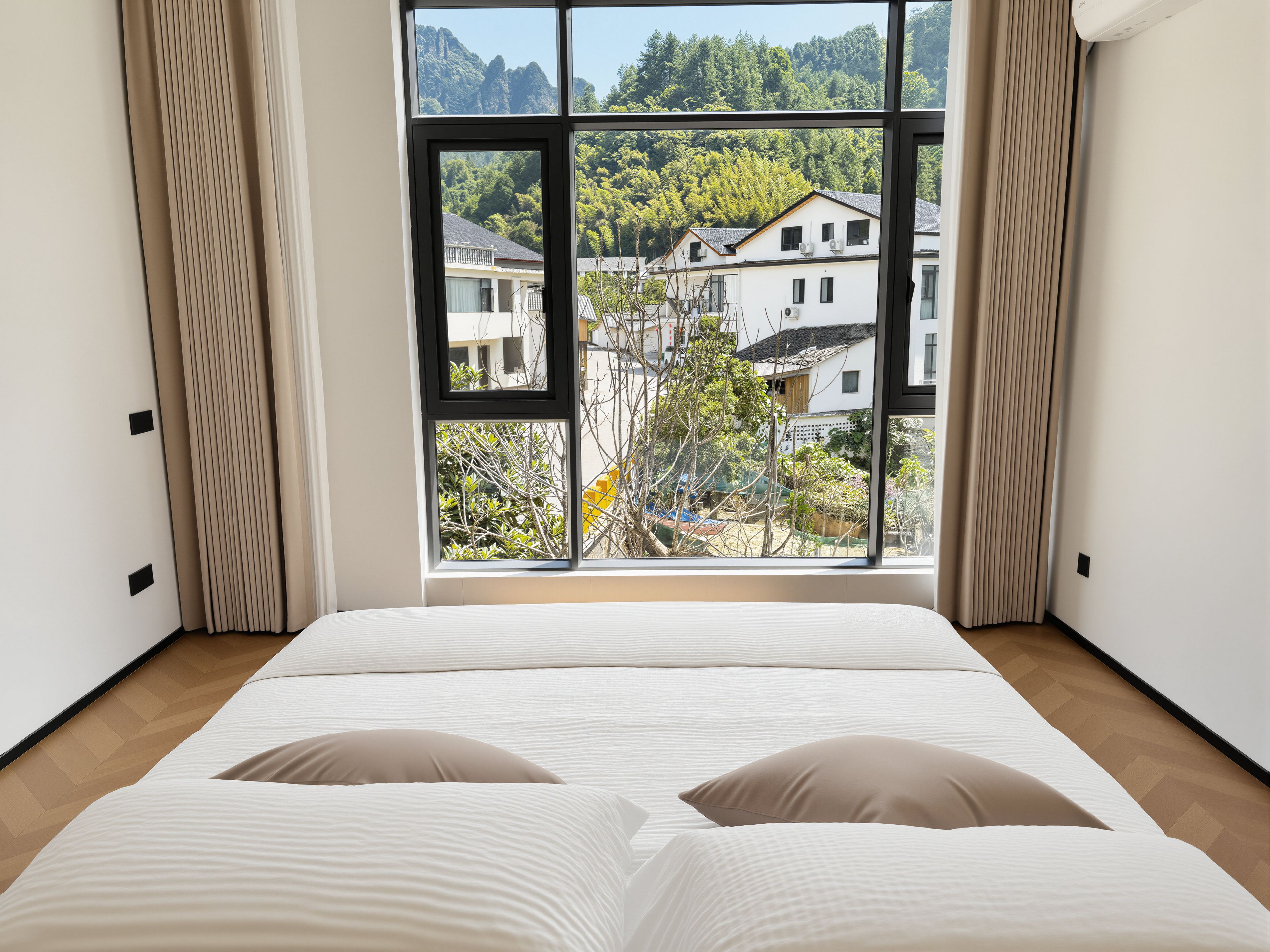 A bedroom with a large window, a bed with two pillows, and a view of a residential area.