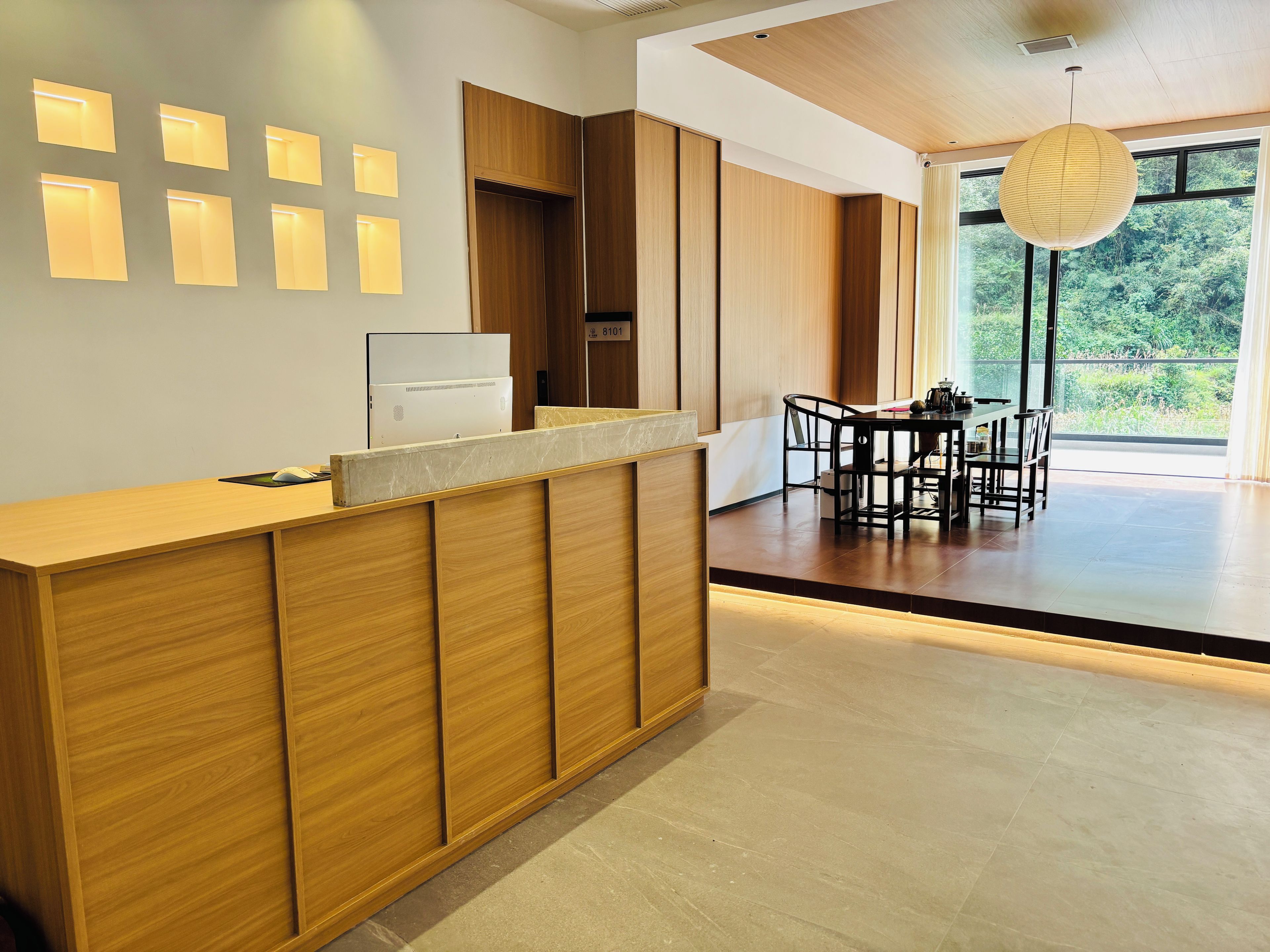 A modern reception desk with a computer, a dining area with a table and chairs, and a view of greenery through large windows.