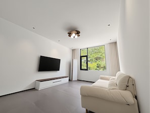 A modern living room with a flat-screen TV mounted on the wall, a sofa, a wooden bench, and a large window with a view of greenery. - Hedui Scenic Homestay - Zhangjiajie (Zhangjiajie)