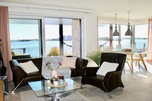 A modern living room with a glass coffee table, leather sofas, and a view of a body of water through large windows. - Holiday apartment with 1 bedroom (Olpenitz)