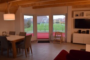 A room with a dining table, chairs, a television, and a view of a garden through a glass door. - Holiday home with 3 bedrooms (Dorum-Neufeld)