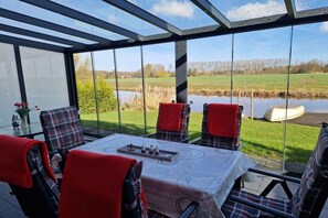 A conservatory with a table and chairs, a chessboard, and a view of a lake and fields. - Holiday apartment with 1 bedroom (Dornum)