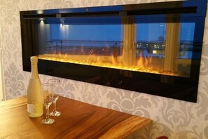 A wall-mounted fireplace with a visible fire, a bottle of white wine, and two wine glasses on a wooden table. - Beach sea view apartment (Borkum)
