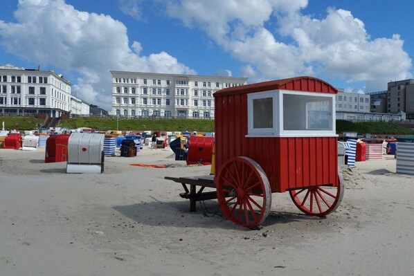A red beach cart with a white window, situated on a sandy beach with buildings in the background. - Beach sea view apartment (Borkum)