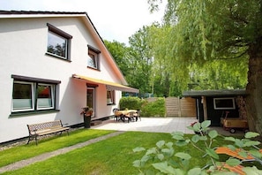 Ein zweigeschossiges Haus mit einer Terrasse, einer Holzbank und einem kleinen Schuppen.