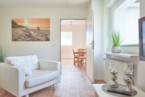 Ein modernes Wohnzimmer mit einem weißen Sofa, einem Bild mit Strandmotiv, einem Essbereich mit Tisch und Stühlen und einem Fensterbrett mit Pflanze.