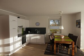 A modern kitchen with white cabinets, a black countertop, and a dining area with a wooden table and chairs. - Borkum sun apartment (Borkum)