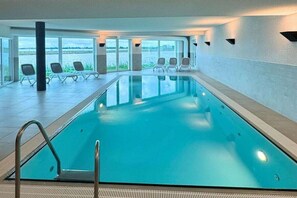 An indoor swimming pool with clear glass walls, a metal ladder, and several lounge chairs. - Holiday apartment with 2 bedrooms (Kappeln)