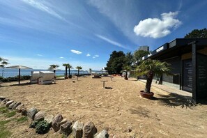 Ett sandigt strandområde med en rad strandstolar och parasoller, en krukväxt i form av en palm och en byggnad med en skylt.
