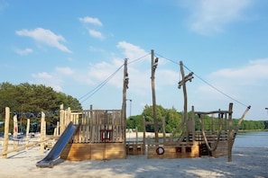 Eine hölzerne Spielkonstruktion mit Rutsche und Kletterelementen, gelegen an einem sandigen Strand mit Bäumen im Hintergrund.
