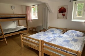 A dormitory room with bunk beds, a small bedside table, a radiator, a window with curtains, and a wall-mounted light switch. - Börstel Abbey - Washhouse (Berge)