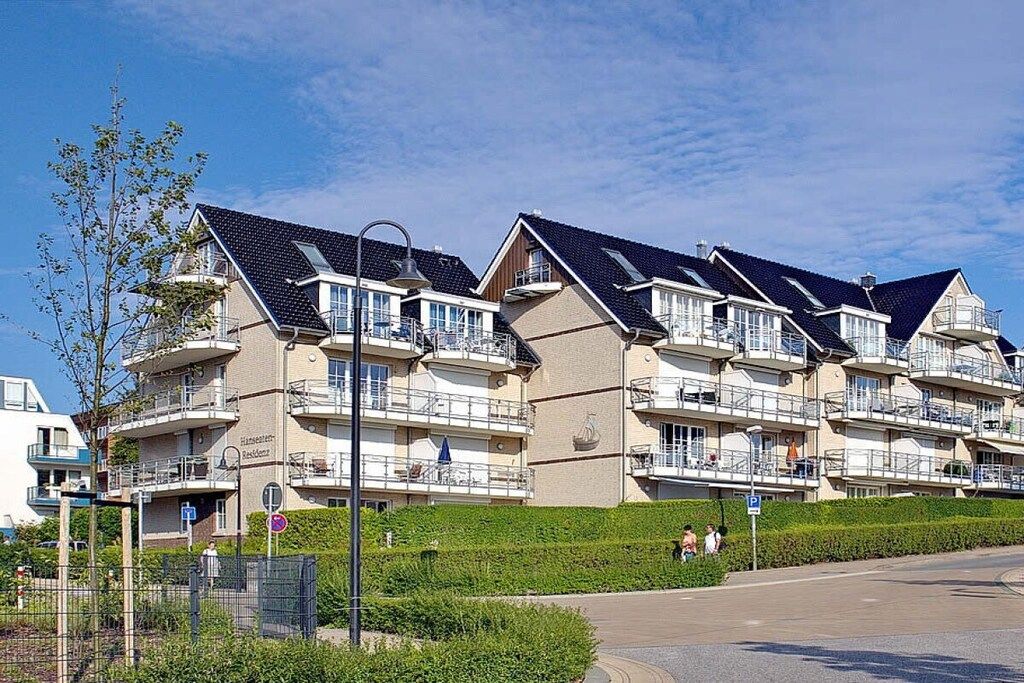 A row of multi-story residential buildings with balconies, a clear sky, and a road in the foreground.