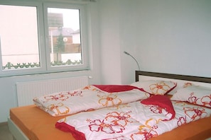 Ein Einzelbett mit Blumenmuster auf der Bettwäsche und einem roten Kissen, ein Fenster mit Blick auf Gebäude und ein Heizkörper unterhalb des Fensters.