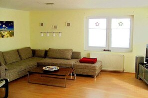 A living room with a sectional sofa, a small ottoman, a coffee table, and a television. There are two windows with curtains, a radiator, and wall-mounted lights. - "Beach apartment Borkum" (Borkum)