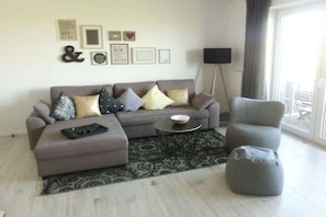 Ein Wohnzimmer mit einer Couch, Hocker, einem Beistelltisch und einem Sessel. An den Wänden befinden sich Dekorationen und es steht eine Stehlampe.