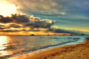 Una platja amb un moll que s'allarga cap a l'aigua, sota un cel clar i un capvespre.