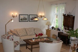 A living room with a sofa, armchairs, a coffee table, and a cabinet. There are two wall-mounted lamps and a window with curtains. - Holiday apartment with 2 bedrooms (Neuenkirchen OT Delmsen)