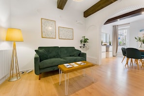 Una sala de estar moderna con un sofá verde, una mesa de centro de madera y una lámpara de pie. Hay dos cuadros en la pared y una planta en maceta sobre una estantería.