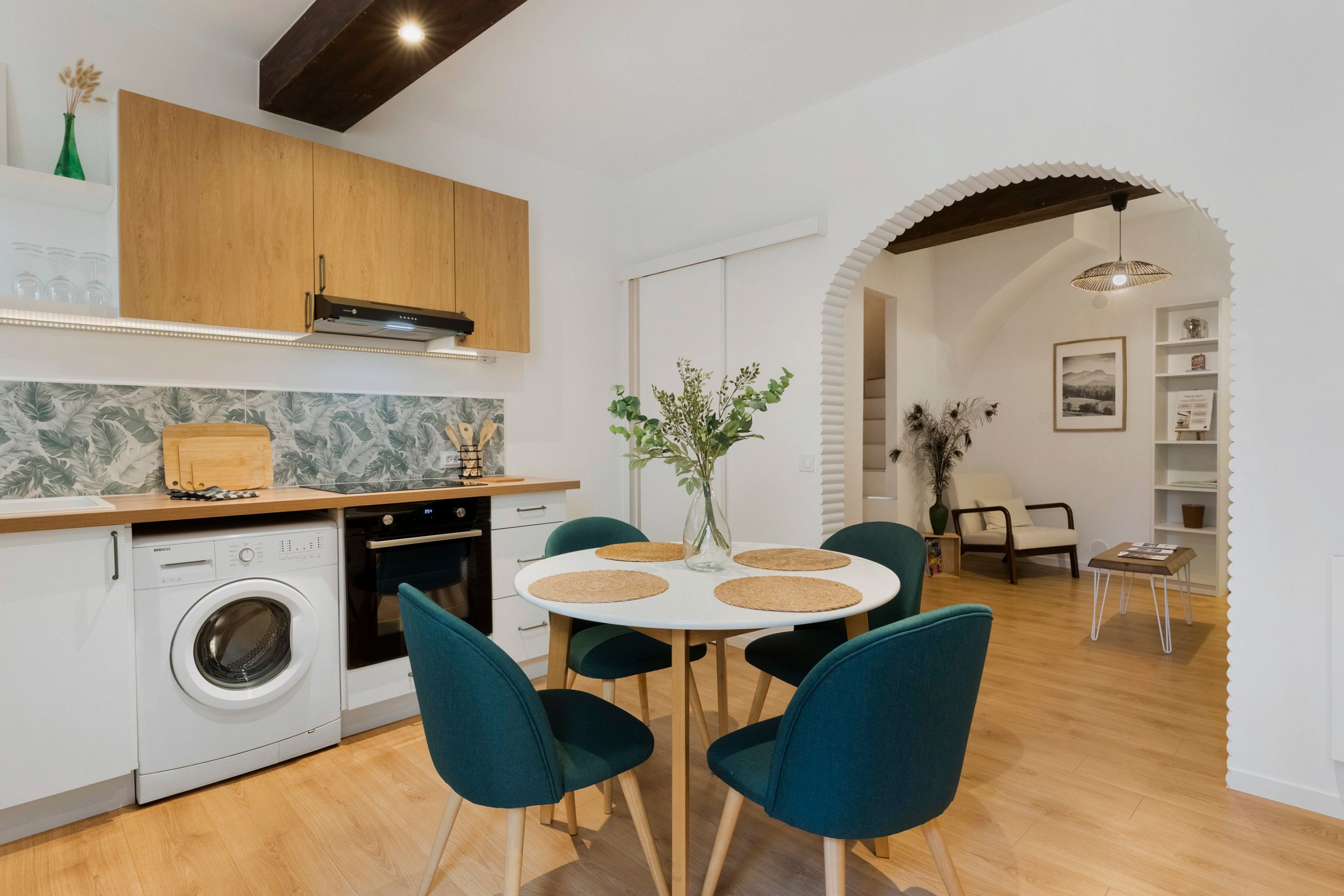 A modern kitchen with a washing machine, a dining area with a round table and chairs, and a living area with a sofa and a lamp.