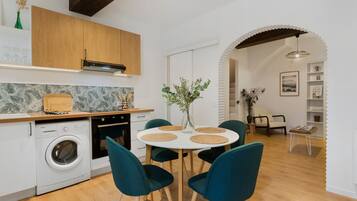 A modern kitchen with a washing machine, a dining area with a round table and chairs, and a living area with a sofa and a lamp.