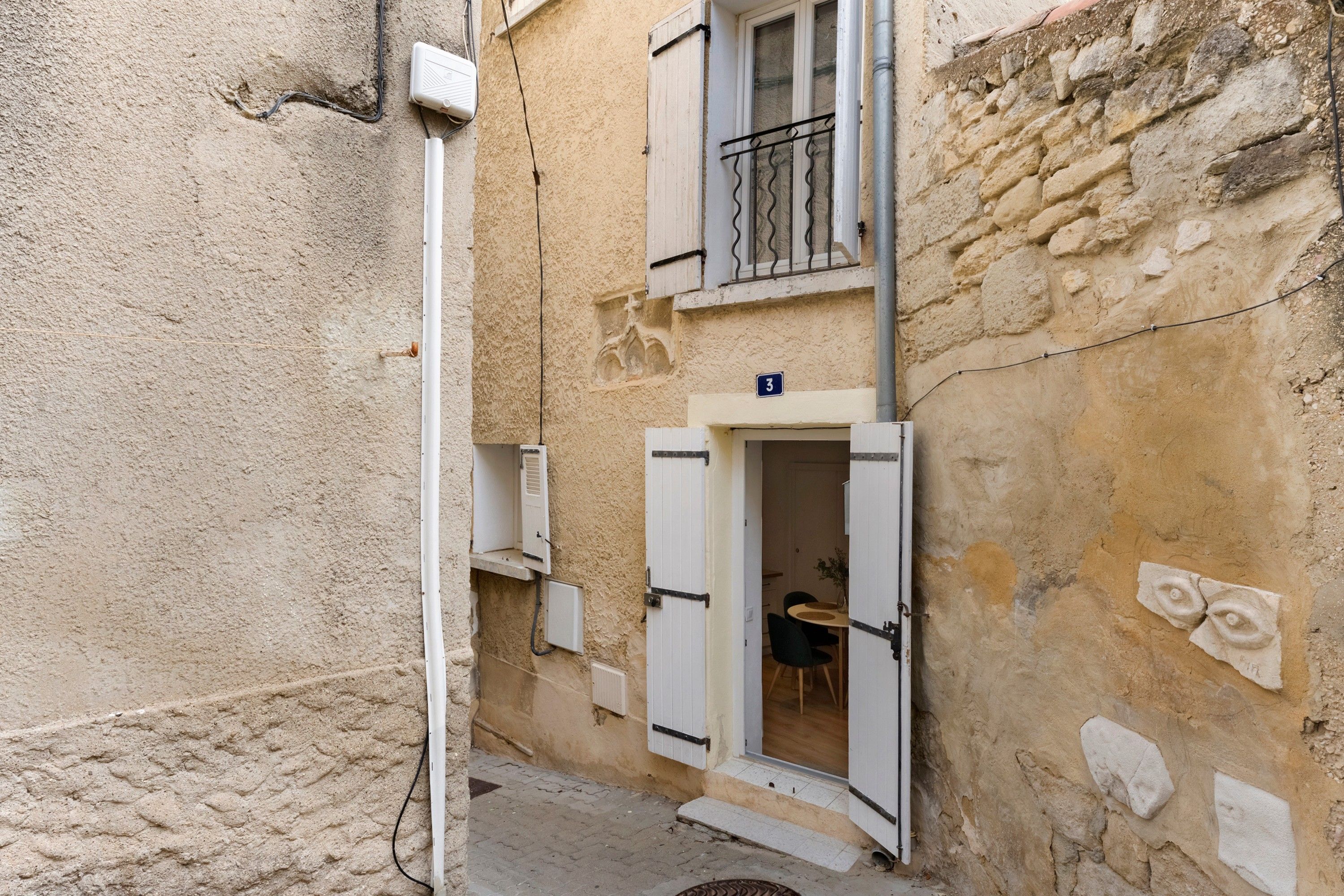 A narrow street with a building featuring a balcony with metal railings and a door leading to a room with a wooden table and chairs.