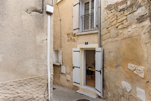 Un carrer estret amb un edifici que té un balcó amb baranes de metall i una porta que dona a una habitació amb una taula i cadires de fusta.