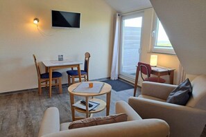 A small, well-lit room with a television mounted on the wall, a dining table with chairs, a sofa, and a small table with a lamp. - Holiday apartment in Juist with swimming pool (Juist)