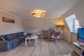 Ein modernes Wohnzimmer mit einem grauen Sofa, einem kleinen Tisch, einem rosafarbenen Sessel und einem Fernseher auf einem Lowboard.