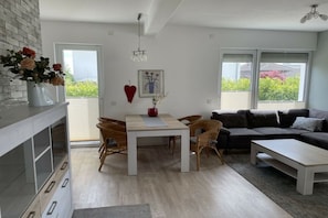 A modern living room with a dining area, a sofa, and a coffee table. There is a fireplace, a window with blinds, and a door leading outside. - Holiday apartment with 1 bedroom (Scharbeutz)