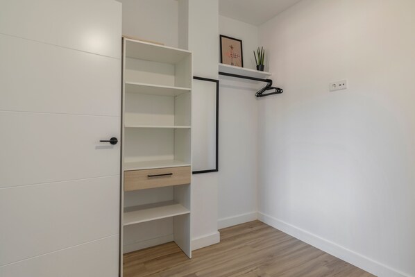 Ein Zimmer mit einem weißen Kleiderschrank, Holzboden, einem schwarzen Kleiderbügel auf einem weißen Regal und einer Steckdose an der Wand.