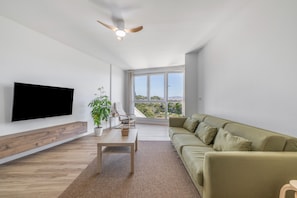 Ein modernes Wohnzimmer mit einem Flachbildfernseher, Holzboden, einem grünen Sofa, einem Deckenventilator und einem großen Fenster mit Blick ins Freie.