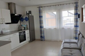 Une cuisine compacte avec des meubles blancs, un four encastré et un réfrigérateur. On y trouve un canapé à rayures et une fenêtre avec des rideaux.