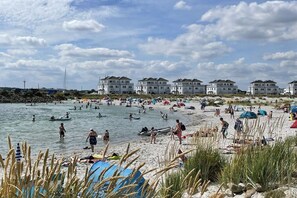 Une plage avec plusieurs bâtiments en arrière-plan, des gens qui nagent et qui prennent des bains de soleil, et des tentes sur le sable.